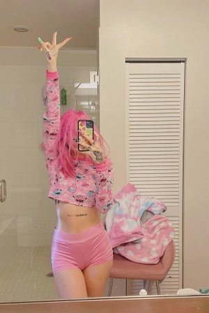 Woman with pink hair in pink crop top and shorts taking mirror selfie in bathroom.