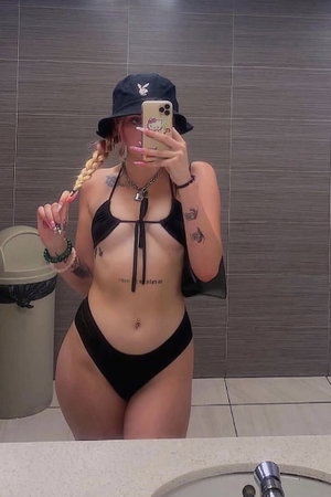 Woman in black bikini and hat taking bathroom mirror selfie.