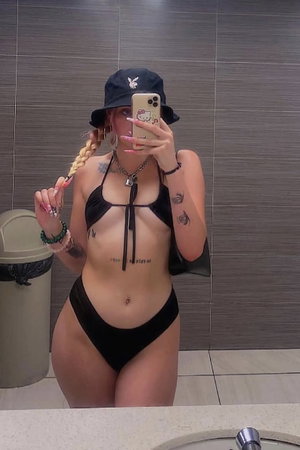 Woman with blonde braid in black bikini taking bathroom mirror selfie.