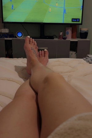 Legs and feet with white toenails on bed, TV in background.