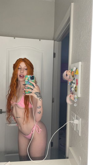 Woman with red hair in pink bikini takes mirror selfie in bathroom.