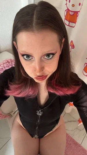 Woman with pink-tipped hair in black zip-up top, sitting indoors near Hello Kitty shower curtain.