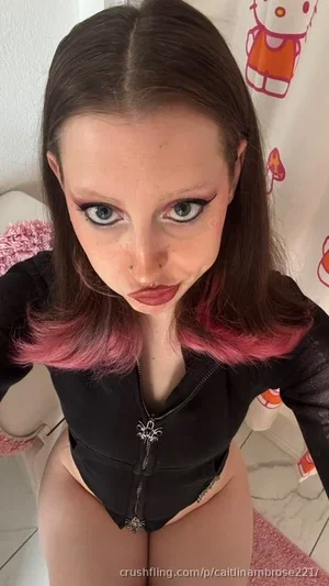 Woman with pink-tipped hair in black zip-up top, posing indoors near Hello Kitty shower curtain.