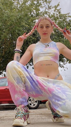 Woman with pink pigtails in crop top and tie-dye pants outdoors.
