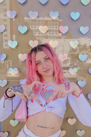 Pink-haired woman in crop top and pants making heart sign against heart wall.