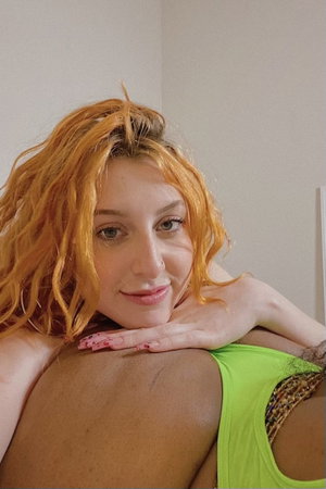 Woman with orange hair in green top resting head on shoulder, indoor setting.