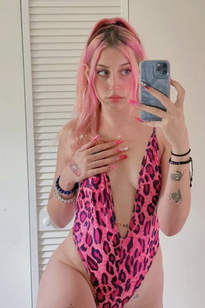 Pink-haired woman in leopard swimsuit taking mirror selfie indoors.
