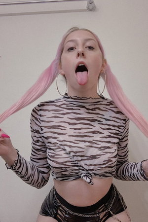 Woman with pink pigtails in zebra-print crop top and black shorts, sticking tongue out indoors.