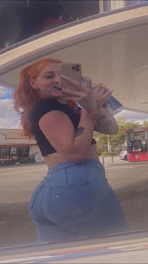 Woman with orange hair in high ponytail, black crop top, blue jeans, taking mirror selfie outdoors.