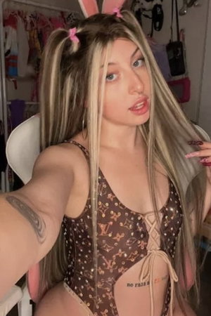 Blonde woman with pigtails in sheer bodysuit taking selfie indoors.