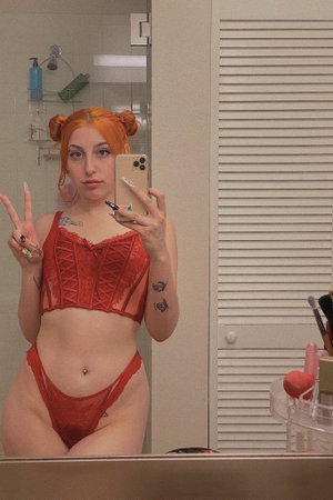 Orange-haired woman in red lingerie taking bathroom mirror selfie.