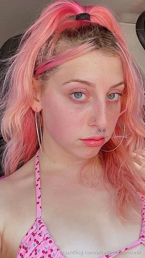 Woman with pink hair and blue eyes wearing a pink cherry-patterned top, indoors.