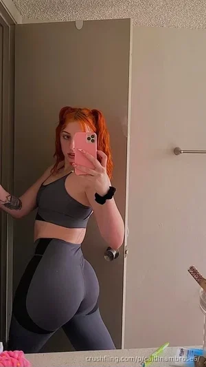 Woman with red hair in pigtails, wearing gray sports bra and leggings, taking mirror selfie in