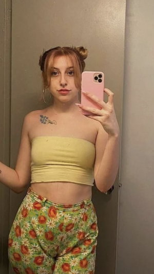 Curvy woman with red hair buns in yellow tube top, floral pants, indoor selfie.