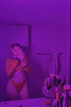 Woman in orange outfit taking mirror selfie in purple-lit bathroom.