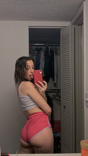 Woman in white crop top and pink shorts taking mirror selfie in bedroom.