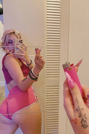 Woman in pink shorts and maroon crop top takes mirror selfie, holding pink lighter.