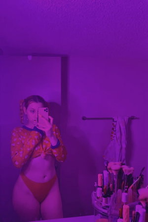 Woman in orange polka-dot top and underwear takes mirror selfie in purple-lit bathroom.