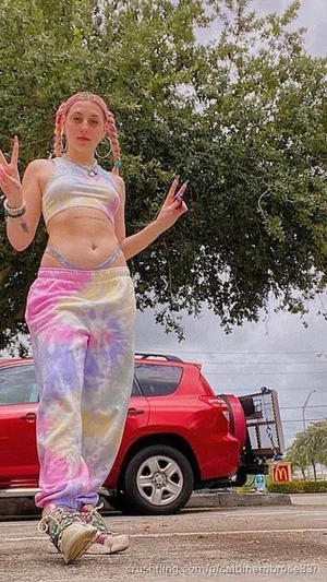 Woman with pink braided hair in tie-dye crop top and sweatpants, standing outdoors near a red car.