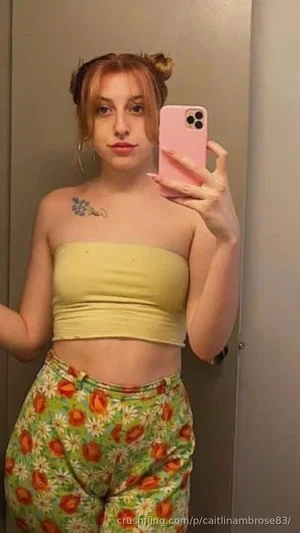 Woman with blonde hair in buns, wearing yellow strapless top and floral pants, taking mirror selfie