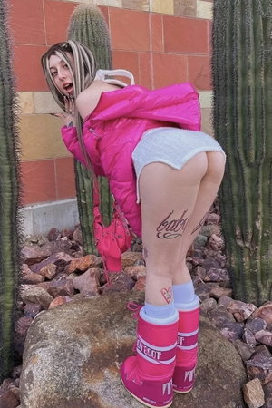 Curvy woman in pink puffer jacket, shorts, tall boots by cacti.