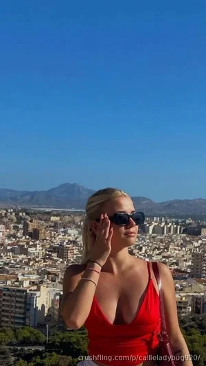 Blonde woman in red top and sunglasses posing outdoors with city and mountains in background.