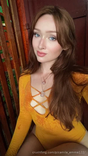 Woman with long brown hair in a yellow lace-up bodysuit, sitting near a wooden fence.
