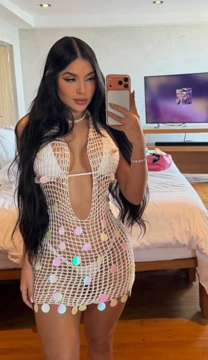 Woman with long dark hair in a sheer mesh dress with colorful dots, taking mirror selfie in bedroom.