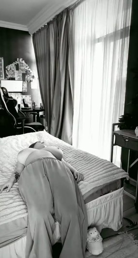 Woman with bare breasts lying on bed, wearing sheer skirt, in bedroom with curtains and desk.