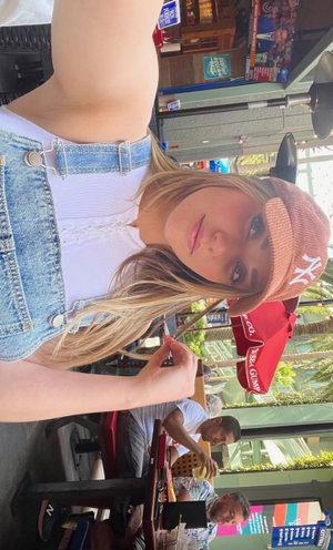 Woman with blonde hair wearing denim overalls and pink cap, posing outdoors at a cafe.