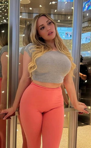 Blonde woman in gray crop top and coral leggings standing in front of mirror in indoor setting.