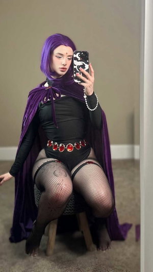 Woman with purple wig in black bodysuit, fishnets, purple cape, indoor selfie.