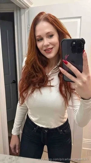 Woman with red hair in white long-sleeve top and dark jeans taking mirror selfie indoors.