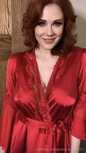 Woman with red hair in a red satin robe with lace trim, smiling indoors.