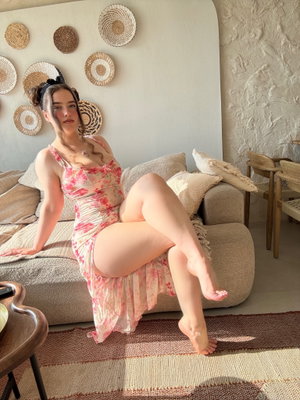 Woman in floral dress sits on couch, legs crossed, in cozy living room with woven wall decor.