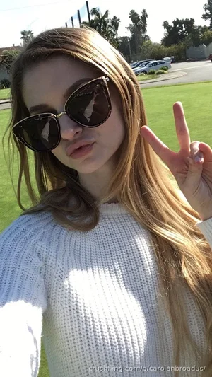 Woman with long hair wearing white sweater and sunglasses, making peace sign outdoors on grass.