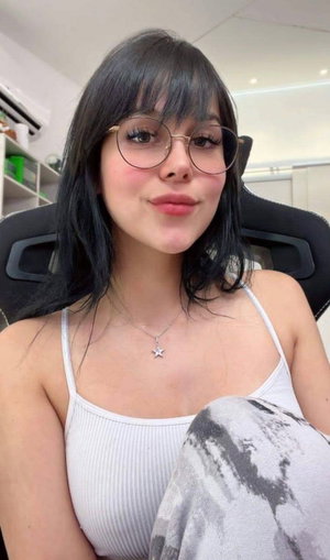 Woman with dark bangs and glasses in white tank top and gray pants on gaming chair.