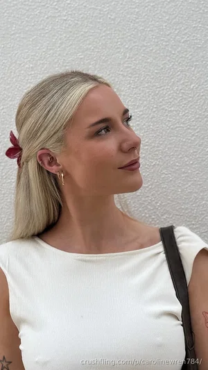 Blonde woman in white top, profile view, standing against textured wall, small tattoos visible.