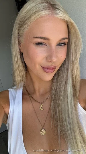 Blonde woman in white tank top with layered necklaces, indoors.