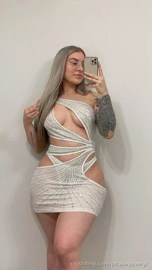 Woman with gray hair and tattoos wears a white cutout dress, taking a mirror selfie indoors.