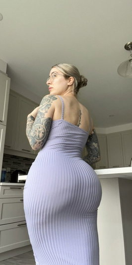 Tattooed blonde woman in purple dress posing in kitchen.
