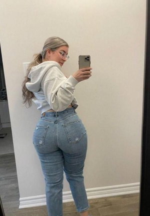 Woman with blonde hair in hoodie and jeans taking mirror selfie indoors.