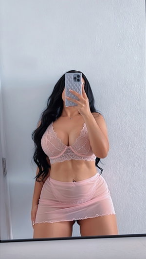 Woman in pink lace bra and sheer skirt taking mirror selfie indoors.