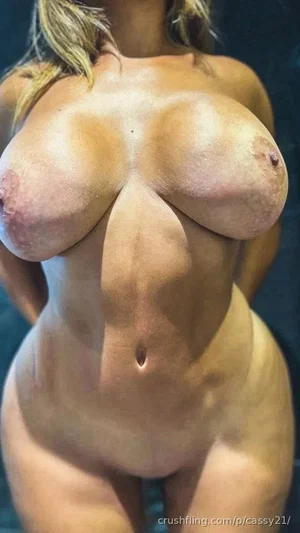 Woman with curvy body, bare chest, standing indoors in dim lighting.