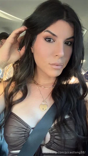Woman with long dark hair in sheer black top, wearing gold necklace, taking mirror selfie indoors.