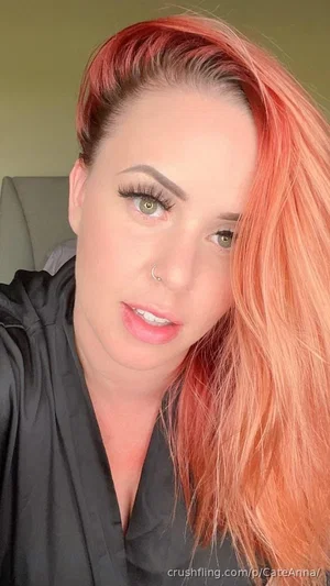 Woman with bright orange hair and green eyes, wearing a black top, taking a close-up selfie indoors.