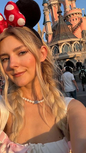 Blonde woman in Minnie Mouse ears and pink dress, posing in front of castle.