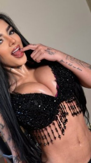 Woman with long black hair and tattoos wears black beaded top, posing indoors.