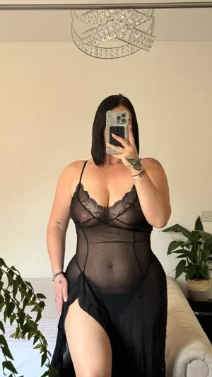 Woman in black sheer lingerie posing in bedroom with plants and chandelier.