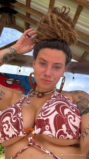 Woman with dreadlocks and tattoos in a patterned bikini top, outdoors under a wooden roof.